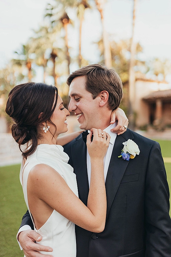 Couple portrait of bride and groom embrace with a forehead touch, her hand on his lapel, palm trees and building behind