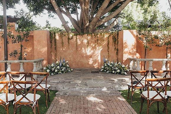 Ceremony setup with outdoor ceremony seating of cross-back chairs and white cushions facing a stone stage in a shaded courtyard walkway