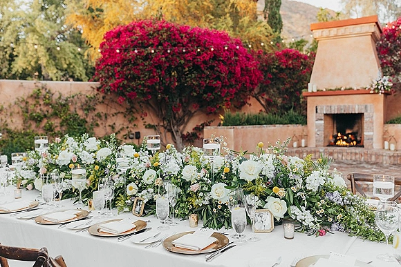 Reception tablescape with wedding table garland, white roses and greenery, bud vases and floating candles under string lights on a patio
