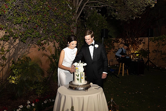 Cake cutting moment as bride in minimalist dress and groom in black tux slice a tall wedding cake on silver stand under string lights outdoors