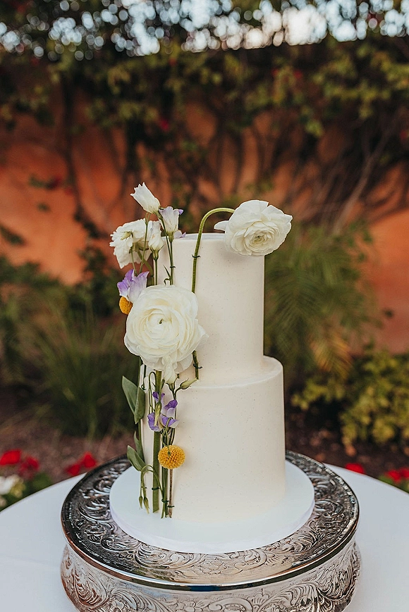 Wedding cake with a floral topper of white ranunculus, purple blooms and yellow billy balls on a silver stand outdoors in a garden