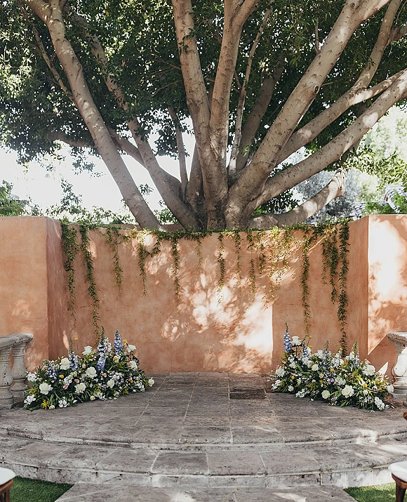 Ceremony backdrop with asymmetrical white and blue floral arrangements and greenery garlands against a stucco wall on a stone terrace