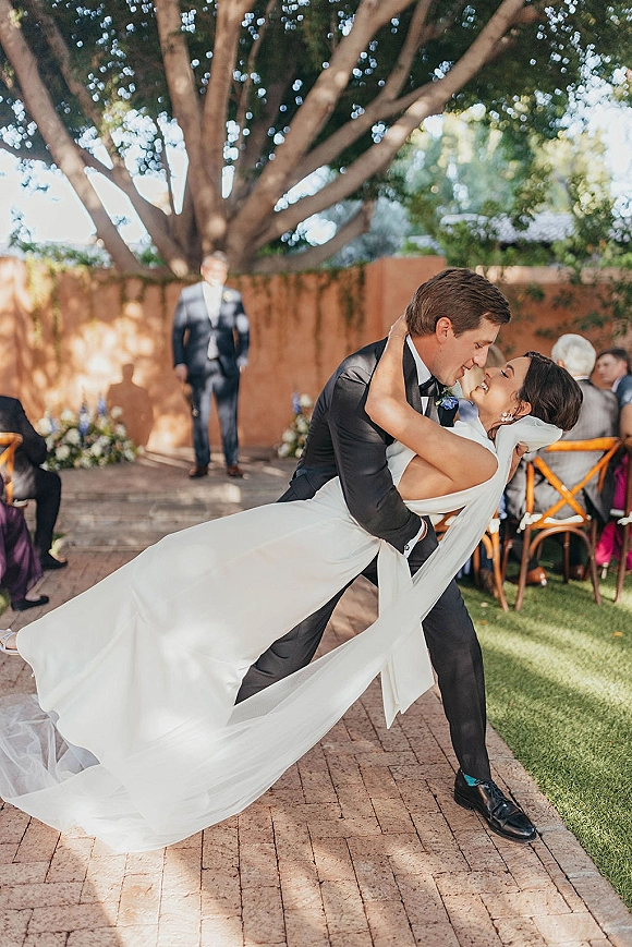 Wedding couple portrait of groom dipping the bride for a kiss, her veil flowing in a courtyard ceremony with brick walkway and guests