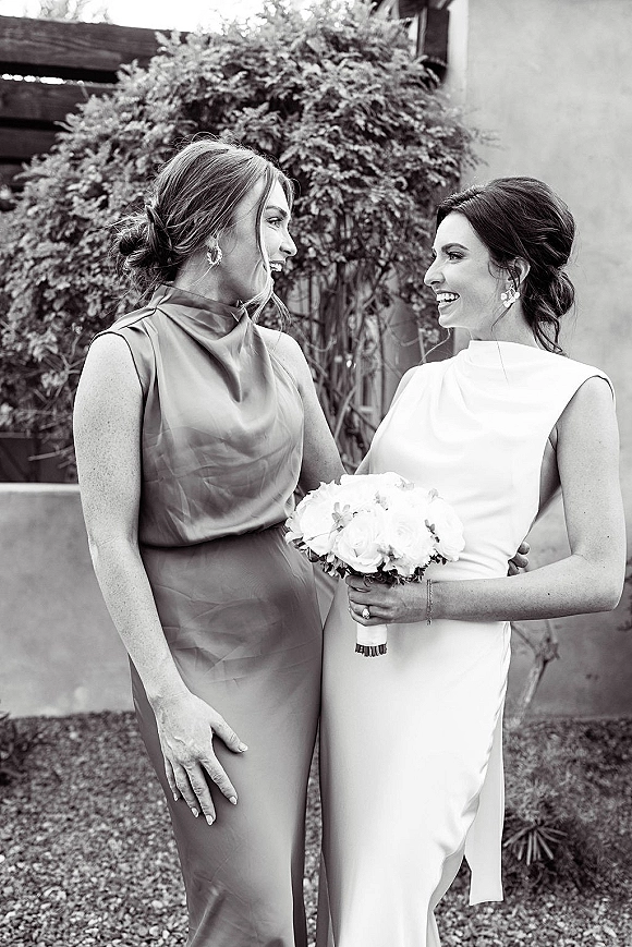 Same sex couple portrait of two brides in modern high neck wedding dresses holding a white rose bouquet, facing each other by a stucco wall