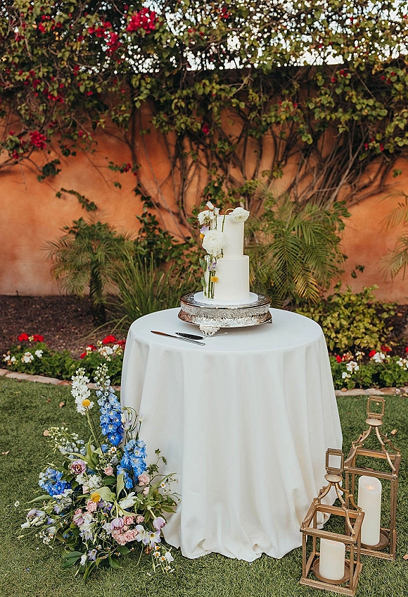 Wedding cake display featuring a two tier wedding cake with floral topper on a silver stand, with lanterns and candles by a vine-covered stucco wall