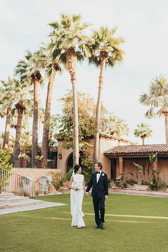 Couple portrait of bride and groom holding hands, bride with white bouquet and groom in black tuxedo by palm-lined villa lawn under blue sky