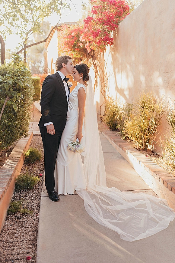 Wedding couple portrait with a forehead kiss, bride in a long veil holding a bouquet beside a sunlit garden walkway and stucco wall