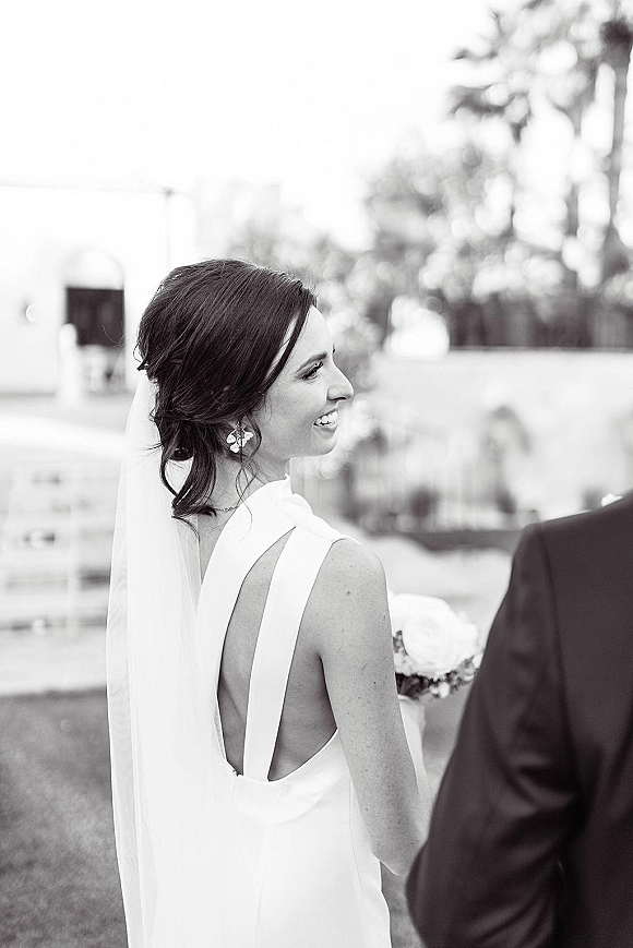 Bridal portrait in black and white, bride in a backless wedding dress with veil and bouquet, looking back on a lawn by trees and a white building