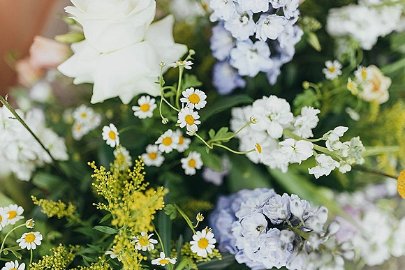 Wedding floral arrangement of white and blue wedding flowers featuring white roses, daisies, blue hydrangea, and greenery against blurred foliage