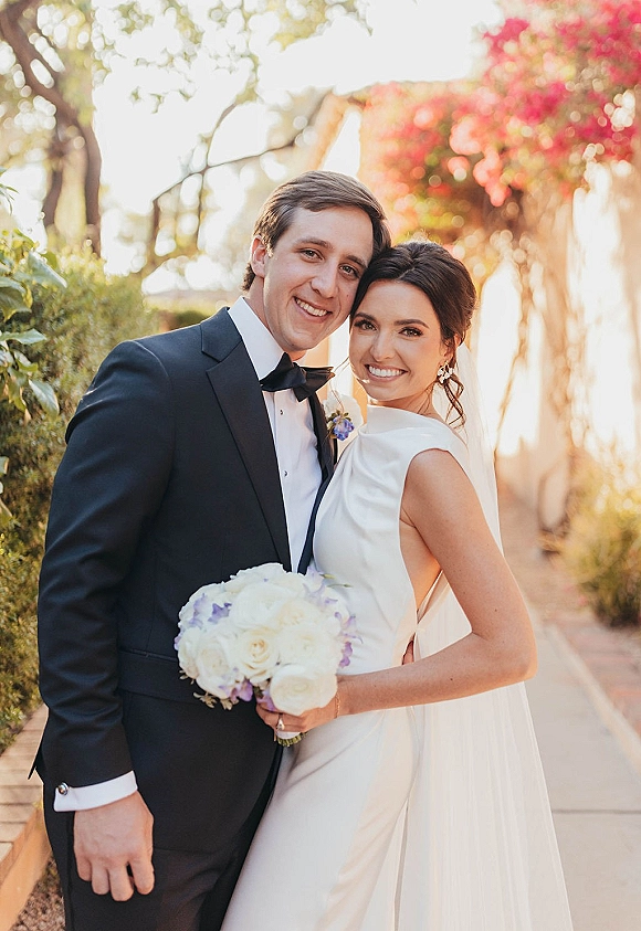 Couple portrait of bride in sleek wedding dress holding a white and lavender bouquet, smiling beside groom in tux on a garden walkway