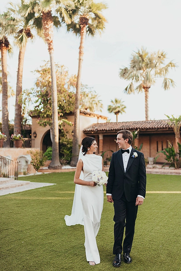 Couple portrait of bride and groom walking hand in hand, her veil and white bouquet flowing in a palm-lined courtyard by stucco building