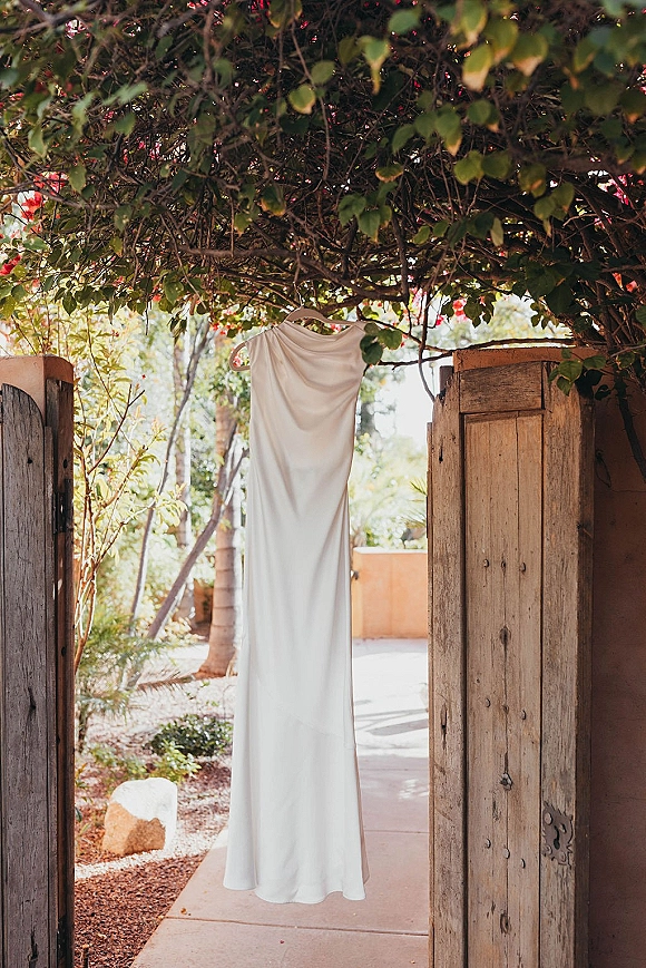 Wedding dress hanging on a hanger, minimalist wedding dress in a rustic wooden doorway beneath a vine canopy on a sunlit garden path