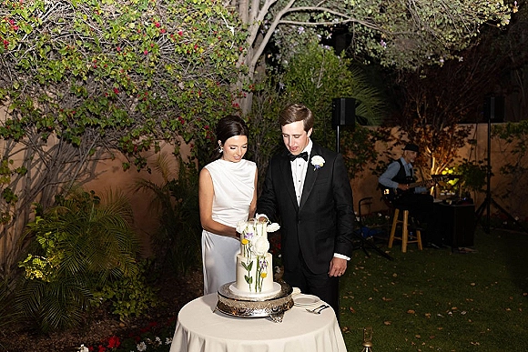 Cake cutting with bride and groom at a white tablecloth table, slicing a white tiered wedding cake with fresh flowers in a garden at night