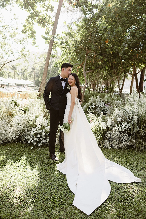 Couple portrait of groom kissing bride’s forehead as she holds a bouquet, her strapless dress with bow train glowing in sunlit garden greenery