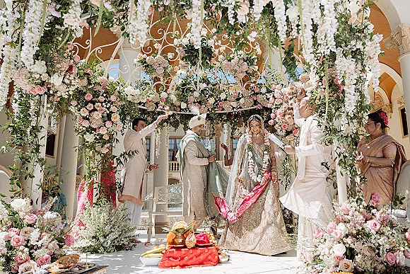 Wedding ceremony moment with a couple in garlands under a floral mandap and sacred fire, set in an arched courtyard with white columns