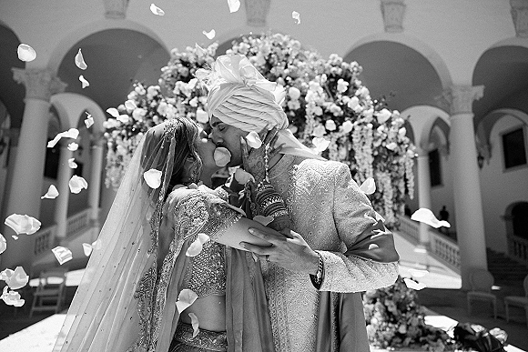 Wedding kiss portrait of newlyweds kissing under a floral arch as petals fall, framed by an arched courtyard with columns and balcony