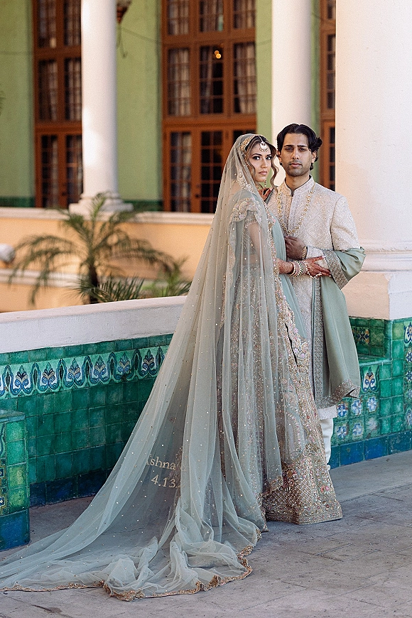 Couple portrait of a South Asian bride in embroidered lehenga and veil with groom in sherwani, posing by columned courtyard doors