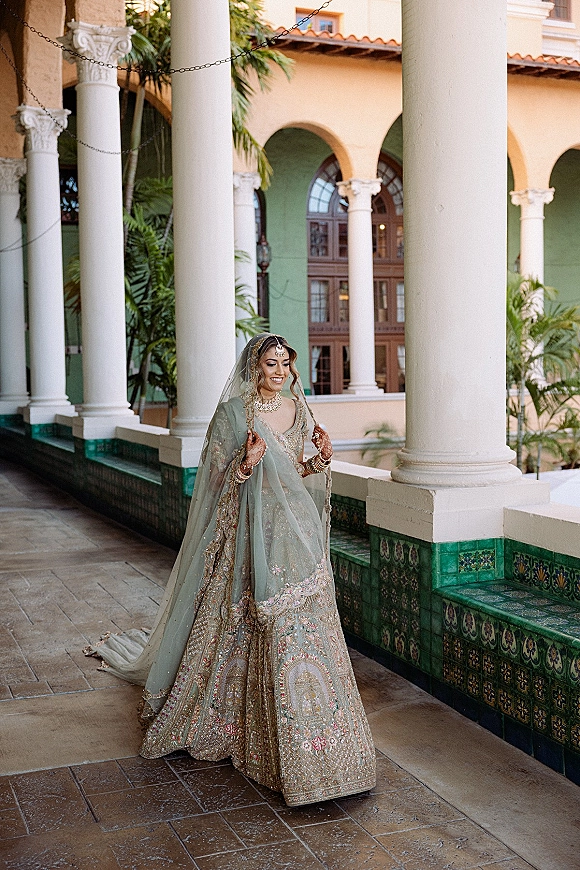 Bridal portrait of a South Asian bride in an embroidered lehenga with veil and jewelry, looking down in a white-column courtyard walkway