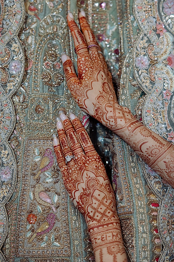 Bridal mehndi hands with henna designs and a pink gemstone engagement ring resting on teal embroidered fabric with sequins