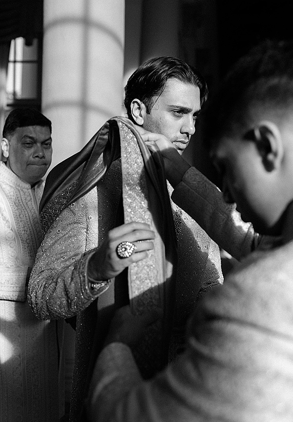 Groom getting ready adjusts an embellished shawl over an embroidered sherwani, statement ring visible in soft window light indoors