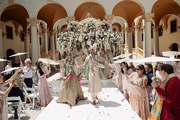 Wedding recessional as bride and groom walk the aisle holding hands while guests toss petals in an arched courtyard with hanging florals