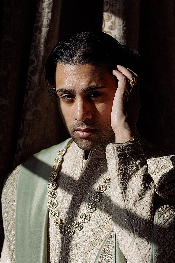 Groom portrait in an ivory embroidered sherwani groom look with a sage green shawl and beaded necklace, lit by window shadows and curtains