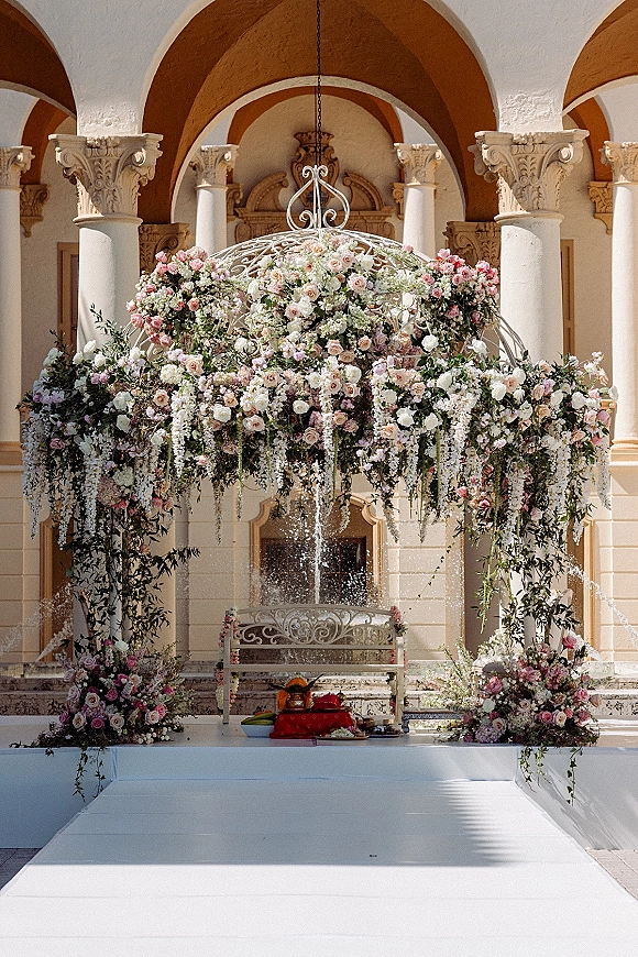 Wedding ceremony backdrop with a hanging floral chandelier of blush and white roses and cascading greenery beneath a white gazebo in a courtyard colonnade