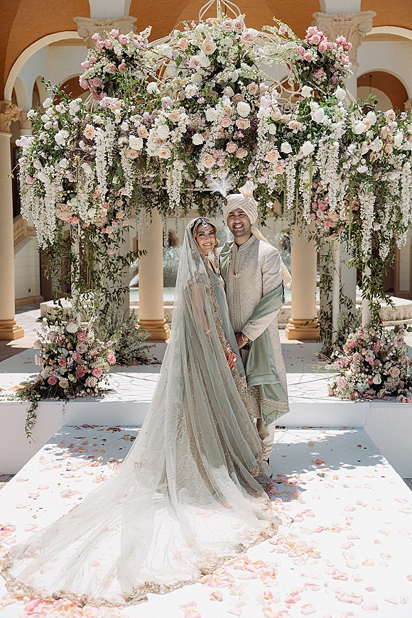 Couple portrait at a hindu wedding mandap, bride in sage lehenga and veil beside groom in turban under hanging floral canopy in a courtyard
