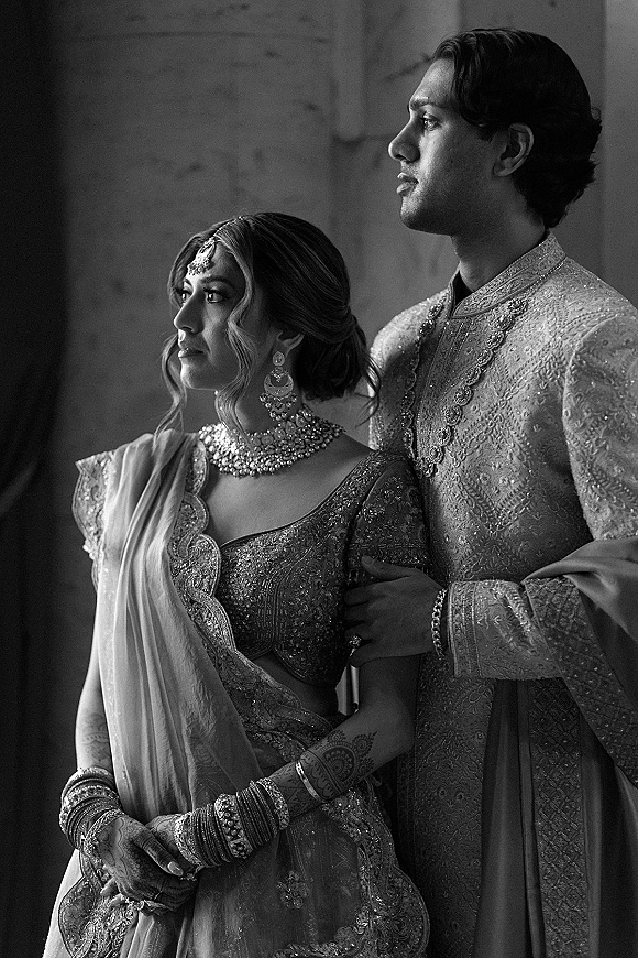 Couple portrait of an Indian wedding couple in lehenga and sherwani, standing close by a wall with moody window light and shadows