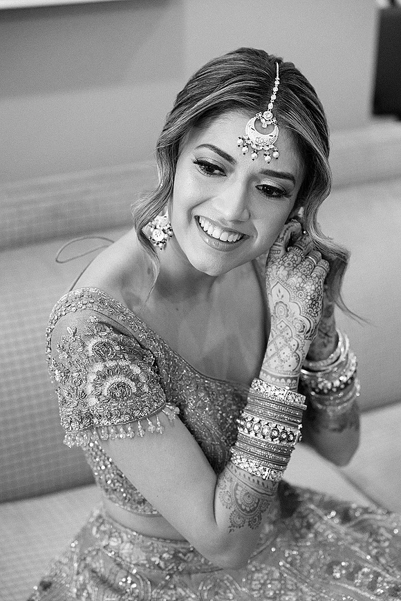Bridal portrait of an Indian bride with henna mehndi on her arms, wearing a maang tikka while seated on a sofa indoors