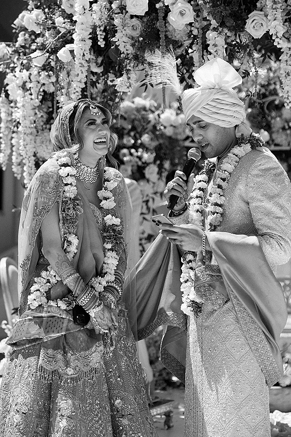Wedding vows as groom reads from phone into microphone while bride laughs beside him, both in garlands before a floral backdrop