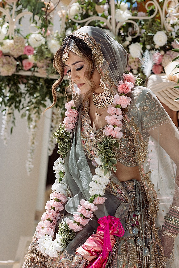 Bridal portrait of a south asian bride in an embroidered lehenga with dupatta veil and floral garland, posed by a garden floral arch
