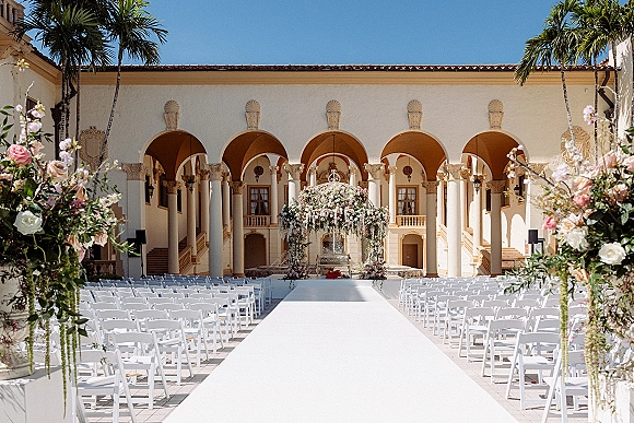 Ceremony setup for outdoor wedding ceremony with white aisle runner leading to a floral arch in an arched courtyard under blue sky