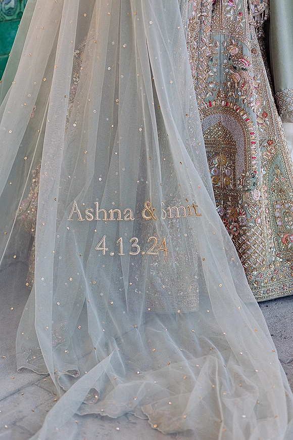 Embroidered wedding veil with personalized bridal veil names and date in script, sequin-dotted tulle over pastel lehenga on stone floor by teal wall