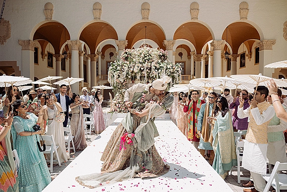 Wedding kiss moment at an indian wedding ceremony as groom dips bride in lehenga under floral mandap, guests in sunlit courtyard with lanterns