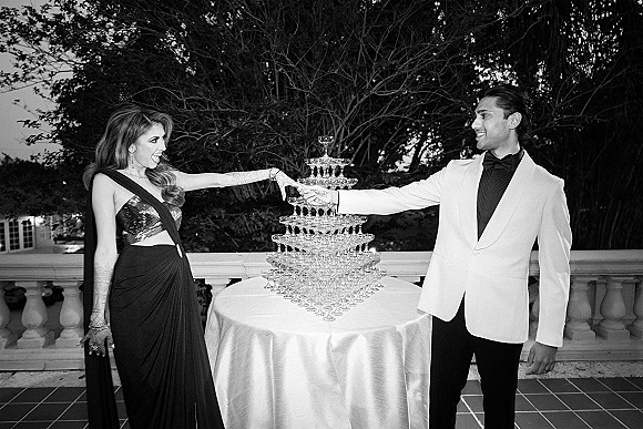 Champagne tower moment as couple in tuxedo jacket and evening gown pours into champagne coupes on a white-clothed terrace table.