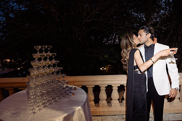 Wedding kiss as the bride and groom kiss beside a champagne tower, on an outdoor terrace under the night sky with trees behind