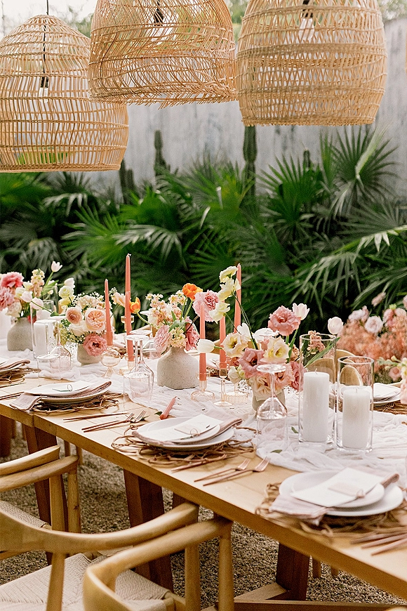 Reception tablescape on an outdoor reception table with rattan chargers, blush napkins, pastel flowers, and candlelight under pendant lanterns amid palms