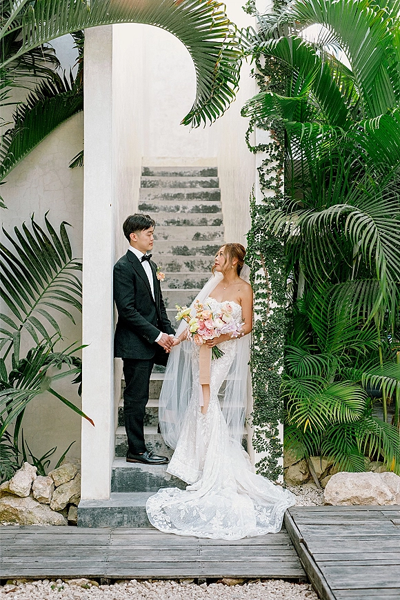 Couple portrait of bride and groom holding hands, her bouquet with long ribbon and veil, under a white stucco arch amid tropical plants