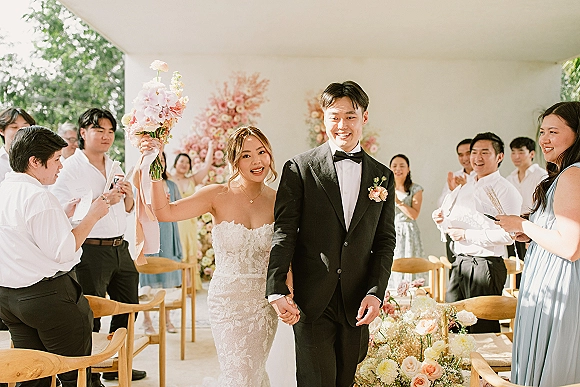 Wedding recessional as bride and groom walk the aisle holding hands, bouquet raised with ribbon streamers while guests cheer on an outdoor patio