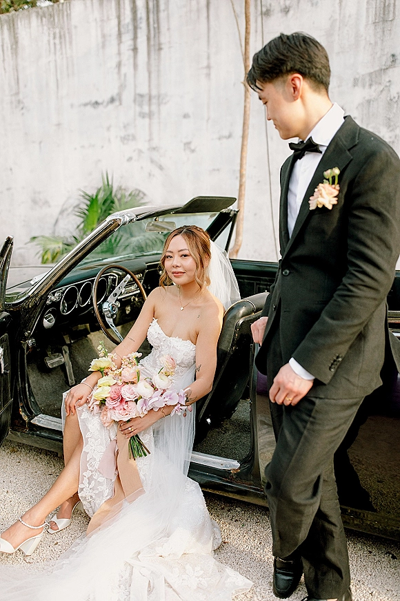 Couple portrait with bride in vintage car holding bouquet as groom stands by convertible, against a stucco wall and gravel drive