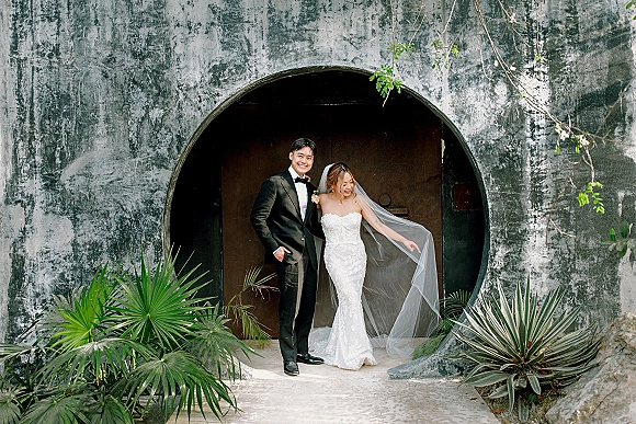 Couple portrait of bride in strapless lace wedding dress and veil with groom in black tuxedo beneath a circular doorway with tropical vines