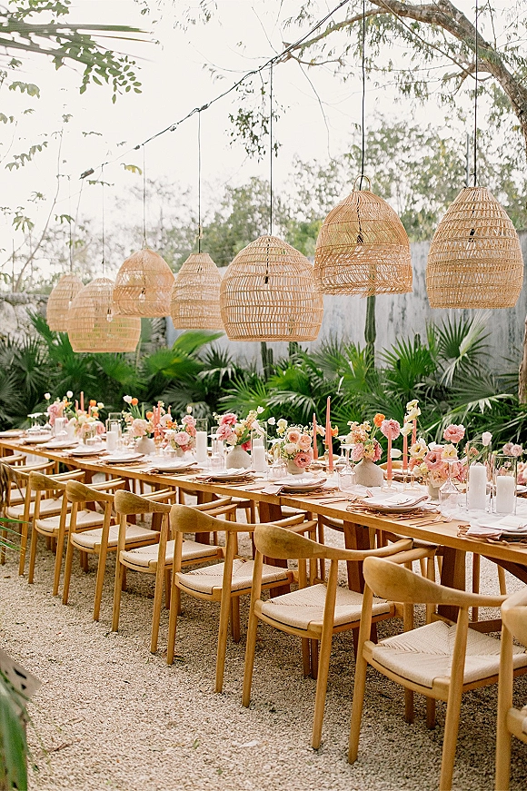 Reception tablescape on an outdoor reception table with wood banquet seating, pastel florals, bud vases, and taper candles under wicker lights