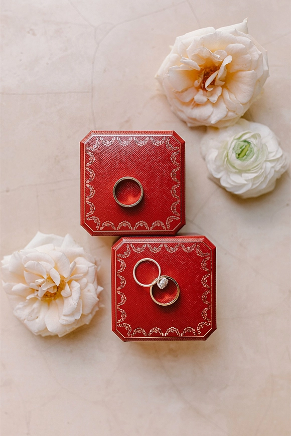 Wedding rings in red ring boxes, a diamond engagement ring and gold wedding bands beside white flowers on a light stone surface