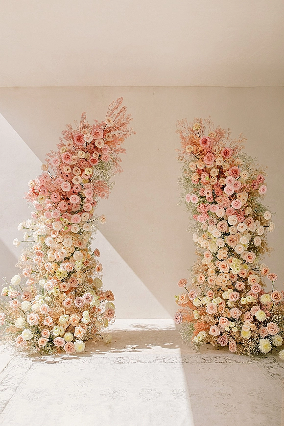 Wedding floral backdrop with pink and blush roses, baby’s breath, and dried stems on floral pillars against a sunlit white wall