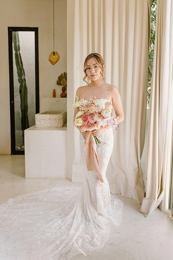 Bridal portrait of bride holding bouquet of roses and orchids in a strapless lace wedding dress with long train, by window light indoors