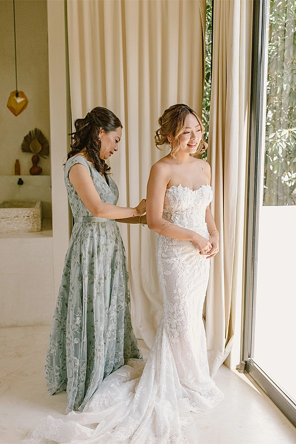 Bride getting ready as a bridesmaid buttons her strapless lace wedding dress by a floor-to-ceiling window with cream curtains