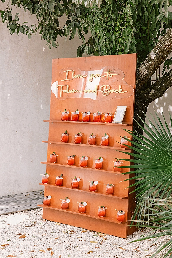 Wedding drink wall with gold-lettered acrylic sign and red cocktails on wooden shelves, set outdoors by a stucco wall and palm fronds
