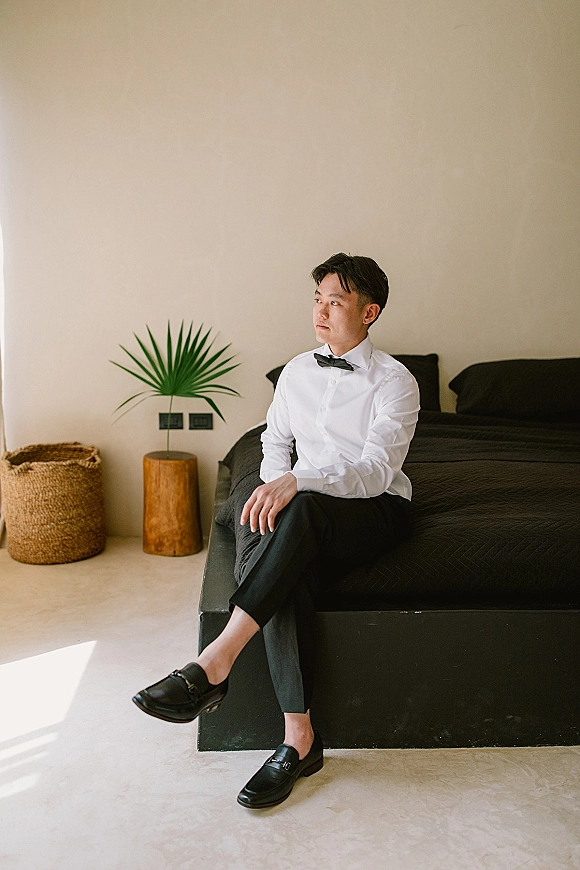 Groom portrait in a white dress shirt and black bow tie, sitting on a bed in soft natural light, beige bedroom backdrop with palm leaf.