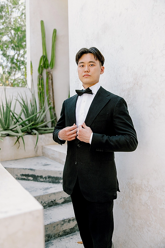 Groom portrait in a black tuxedo and bow tie, standing on concrete steps by a white stucco wall with cactus and greenery